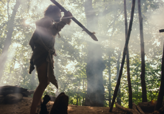 Les hommes du Paléolithique Reconstitution paléolithique : une personne vêtue d'une tenue ancienne, tenant une lance, dans une forêt baignée de lumière matinale